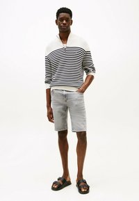 Gestreepte trui in marineblauw en wit, met een ritssluiting bij de kraag, gecombineerd met lichtgrijze denim shorts en zwarte sandalen met een gespdetail.