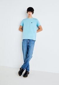 Young man leaning against white wall, wearing light blue t-shirt, blue jeans, and black boots, with arms behind his back.