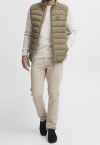 Khakifarbenes Steppwesten mit horizontaler Quiltung und Logo-Patch, getragen über einem hellen Pullover. Beigefarbene Hose und schwarze Schuhe vervollständigen das Outfit.