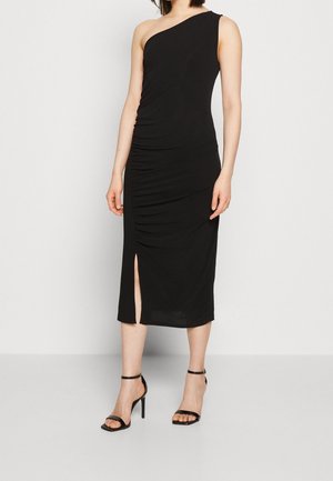 Femme portant une robe midi noire à une épaule avec une fente sur le côté et des sandales à talons ouverts noires, se tenant contre un fond uni.