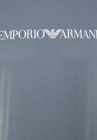 Harmaa puuvillainen t-paita, jossa on suuri valkoinen "Emporio Armani" -logo tyyliteltyine siipineen. Lyhyet hihat, joissa on tasainen ja sileä pinta.