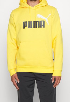 Mężczyzna w jasnej żółtej bluzie Puma z kapturem, z kieszenią z przodu i czarnym logo Puma, w połączeniu z czarnymi spodniami Puma.