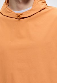 Κοντινό πλάνο ενός ατόμου που φοράει ανοιχτό πορτοκαλί φούτερ με κουκούλα και λευκό μπλουζάκι ορατό στο γιακά.