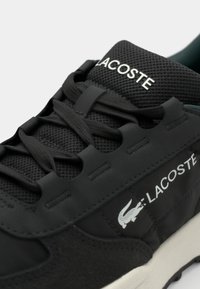 Baskets Lacoste noires en mesh et suède, dotées de lacets noirs et du texte Lacoste blanc ainsi que du logo crocodile sur le côté et la languette.