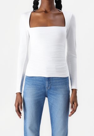 Nainen, jolla on valkoinen pitkähihainen yläosa, jossa on neliömäinen pääntie, ja vaaleansiniset farkut, seisoo yksinkertaista valkoista taustaa vasten.