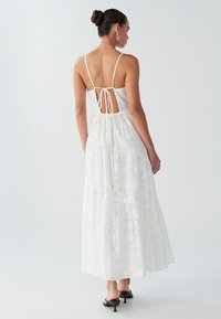Robe maxi en coton blanc avec bretelles ajustables, dos ouvert et design à étages avec motifs à œillets. Texture lisse et légère.