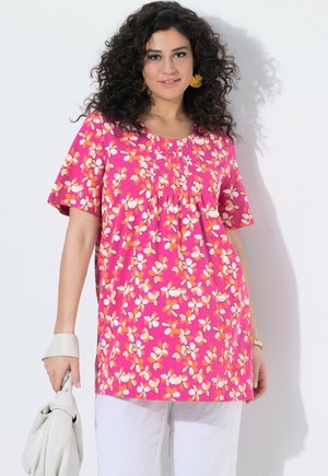 Kobieta z kręconymi włosami w różowej, kwiecistej bluzce z krótkim rękawem i białych spodniach, trzyma białą torebkę, stoi na tle białej ściany.