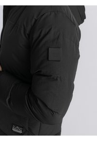 Chaqueta acolchada negra con una superficie brillante y texturada; cuenta con un pequeño parche de logo negro en la parte superior del brazo. Cierre frontal con cremallera.
