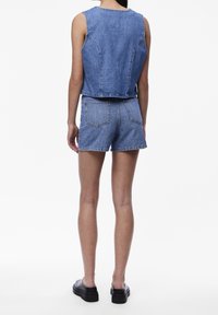 Vrouw staande met de rug naar de camera, gekleed in een mouwloos denimvest, denim shorts met achterzakken en zwarte plateauzolen, op een witte achtergrond.