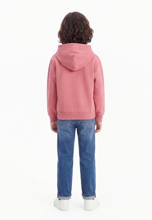 Enfant aux cheveux bruns bouclés portant un sweat à capuche rose, un jean bleu et des baskets blanches, debout de dos sur un fond blanc.