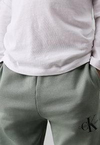 Camicia a maniche lunghe grigia chiara in cotone abbinata a pantaloni tuta verdi con texture morbida, cintura elastica e dettaglio logo nero.