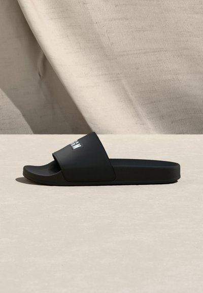 Sandale à glissière noire avec une large bride portant un texte blanc, posée sur une surface claire avec un fond en tissu beige.