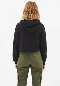 Svart kort hoodie med dragsko, gjord av mjukt tyg, ihop med löst sittande olivgröna byxor med sidfickor.