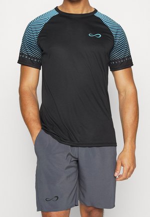 Mann trägt schwarzes und blaues kurzärmeliges Sportshirt mit gestreiften Ärmeln und graue Shorts mit schwarzem Logo, steht mit einer Hand in der Tasche.