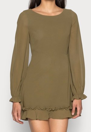 Frau trägt ein olivgrünes, langärmliges Kleid mit Rüschen an den Ärmelabschlüssen und am Saum, steht vor einem einfarbigen hellen Hintergrund.