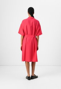 Bringebærfarget omslagskjole med korte ermer, belted liv og knelengde. Mykt, teksturert stoff med minimale designdetaljer.