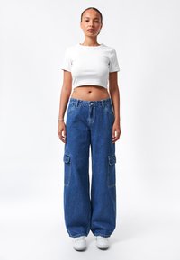Camiseta blanca corta con mangas cortas combinada con pantalones cargo azules de mezclilla holgados que presentan bolsillos laterales. El modelo lleva zapatillas blancas.