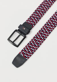 Υφασμένη ζώνη με πλεγμένο σχέδιο σε κόκκινο, λευκό και σκούρο μπλε, μαύρη μεταλλική αγκράφα, και μαύρο δερμάτινο άκρο και βρόχο με ένα μικρό έμβλημα σημαίας.