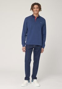 Polo a maniche lunghe blu navy con colletto rosso e logo ricamato, abbinato a jeans blu scuro e sneakers bianche con accenti colorati.