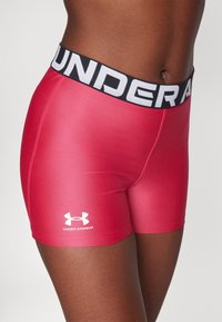 Červené sportovní šortky z hladké tkaniny, s širokým černým pasem s nápisem "UNDER ARMOUR" v bílém písmu a bílým logem jako doplňkem.