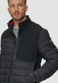 Zwarte pufferjack met textuur nylon schouders, hoge kraag, ritssluiting aan de voorkant en borstzak. Bevat rode accenten op de binnenkant van de kraag.