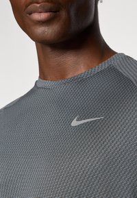 Sivý atletický tričko s horizontálnym pruhovaným vzorom, okrúhlym výstrihom a malou reflektívnou logom Nike na hrudi. Hladká, elastická látka.