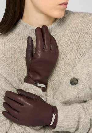 Hnědé kožené rukavice s jemným leskem, s přiléhavým střihem a stříbrnými kovovými doplňky, položené na světle šedém pleteném svetru.