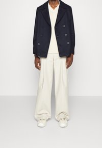 Abrigo cruzado azul marino, suéter beige y pantalones crema de corte holgado; combinados con zapatillas blancas. Texturas suaves y acentos mínimos.