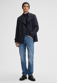 Cappotto peacoat navy con design doppiopetto, maglione nero a girocollo, jeans dritti azzurri chiari e scarpe in pelle nera. Tessuto in lana.