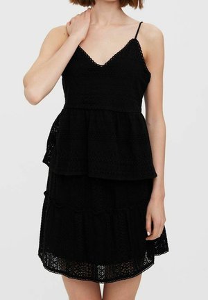 Vrouw die een zwarte mouwloze kanten jurk draagt met een gelaagde rok en dunne bandjes, haar hand rustend op haar nek, tegen een effen lichte achtergrond.