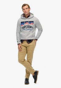 Felpa grigia con cappuccio, decorata con una grafica di montagne e la scritta "SUPERDRY OUTDOOR", indossata sopra una camicia a quadri rossa, abbinata a pantaloni kaki e scarpe nere.