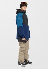 Blå og sort snejakke med hætte, beige cargo-bukser og sorte sneakers. Jakken har farvet blokserie og praktiske lommer.