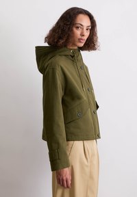 Oliwkowa kurtka z kapturem o skróconym kroju, z dużymi guzikami, przednimi kieszeniami oraz ściągaczem przy szyi.