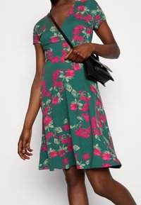 Grön blommig klänning med korta ärmar, med rosa och ljusgröna blommor, A-linjeformad och med en mjuk textur. Åtföljd av en liten svart väska.