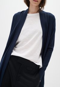 Cárdigan abierto azul marino sobre una camiseta blanca ligera, acompañado de pantalones negros, destacando un ajuste relajado y una textura suave.