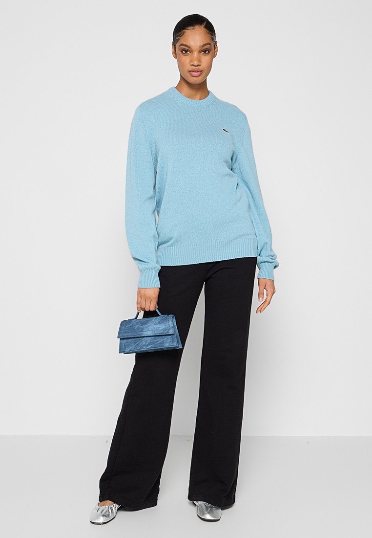 Femme portant un pull bleu clair, un pantalon large noir, des chaussures argentées, tenant un petit sac à main bleu, se tenant contre un fond blanc uni.