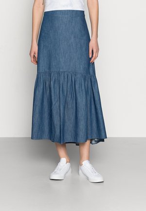 Vrouw draagt een blauwe, gelaagde denim midi-rok gecombineerd met witte sneakers en een wit topje, staand tegen een effen achtergrond.
