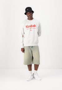 Γκρίζα φούτερ με κόκκινη εκτύπωση "Kodak", ανοιχτό μπλε τζιν σορτς, λευκά αθλητικά παπούτσια και μαύρο καπέλο bucket. Απλός, καλοκαιρινός, καθημερινός στυλ.