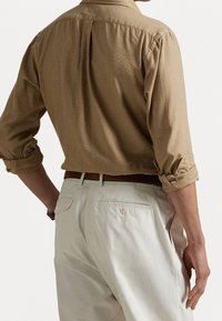 Camicia marrone chiaro con maniche arrotolate, dettaglio in piega sul retro, abbinata a pantaloni beige chiaro con tasca posteriore a bottone.