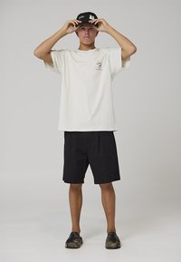 T-shirt bianca in cotone con grafica, pantaloncini neri e cappellino nero. Taglio corto, scollatura ampia e scarpe casual slip-on con motivo mimetico.