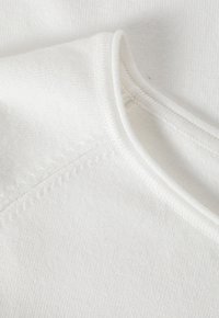 Weißer Baumwollstoff mit glatter Textur, ausgestattet mit einem runden Ausschnitt und sichtbaren Nahtdetails entlang der Nähte.