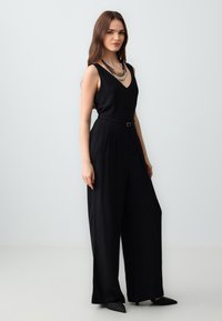 Zwarte jumpsuit met V-hals, wijde pijpen, een ceintuur in de taille en lichtgewicht stof. Accentueert met een gelaagde metalen ketting.