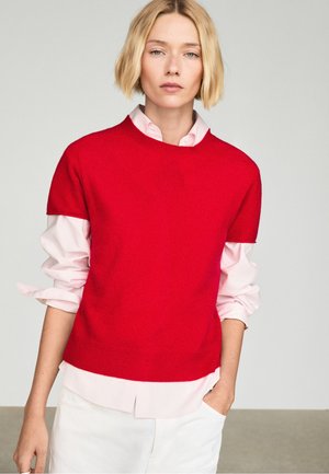Blond kvinde med kort hår har en knald rød kortærmet sweater over en lys rosa skjorte med knapper og optrukne ærmer samt hvide bukser.