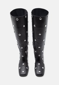 Μαύρες δερμάτινες μπότες μέχρι το γόνατο με τετράγωνα δάχτυλα, διακοσμημένες με ασημένια καρφιά τοποθετημένα σε διάσπαρτο μοτίβο κατά μήκος της επιφάνειας. Περιλαμβάνει φερμουάρ.