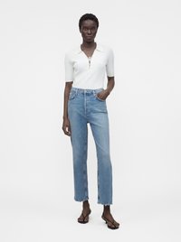 Woman standing wearing a white short-sleeve knit top with lace-up neckline, light blue high-waisted straight jeans, and black open-toe sandals.