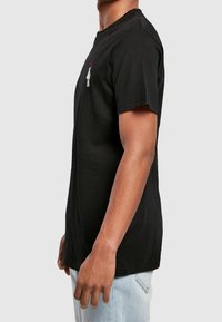 Schwarzes Baumwoll-T-Shirt mit Rundhalsausschnitt, kurzen Ärmeln und einem kleinen Grafikaufdruck auf der Brust. Das Model trägt hellblaue Jeans.