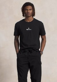 Nuori mies, jolla on dreads-kampaus, pukeutuneena mustaan Ralph Lauren -t-paitaan ja mustiin cargo-housuihin, seisoo kädet taskuissa neutraalia taustaa vasten.