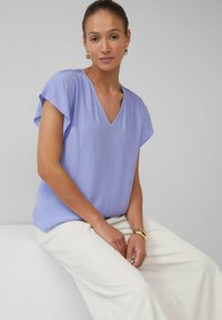 Licht lavendelkleurige blouse met korte mouwen, een V-hals en geplooide schouderdetails. Gekozen in combinatie met een witte broek, gemaakt van soepel materiaal en met een relaxte pasvorm.