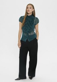 Teal sheer blouse features a high ruffled neck and short sleeves, paired with loose black trousers and pointed black shoes.