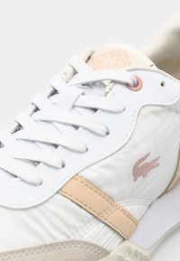 Baskets blanches avec accents beiges, lacets blancs et un petit logo crocodile rose sur le côté près de la cheville.